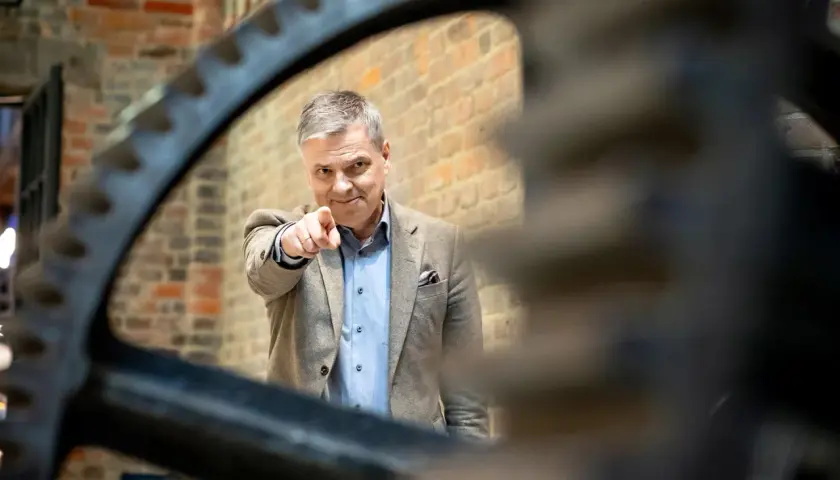 Mężczyzna w garniturze wskazuje palcem, jakby prezentował zestawienie finansowe. Za nim ceglana ściana i zębate koło.