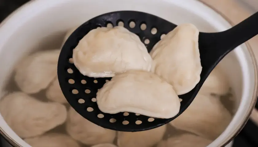 Gotowane pierogi na czarnej łyżce cedzakowej w garnku z wodą.