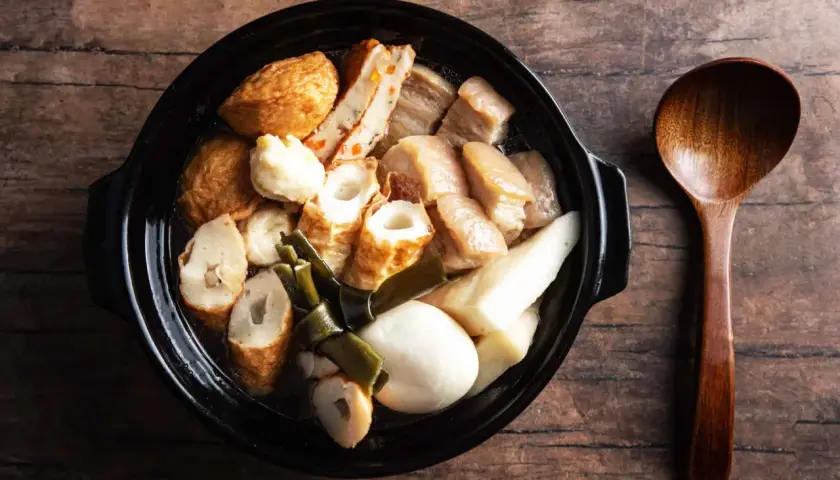 A steaming pot of oden, a Japanese fish cake stew, with various ingredients like daikon and pork belly, served with a wooden spoon.