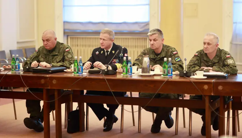 Czterech mężczyzn w mundurach siedzi przy drewnianym stole, przed nimi butelki wody i filiżanki.