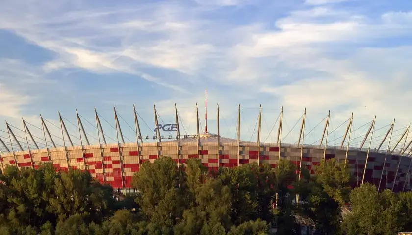 Stadion Narodowy w Warszawie, z imponującą pojemnością, otoczony zielenią, na tle błękitnego nieba z chmurami. Ludzie odpoczywają na plaży.