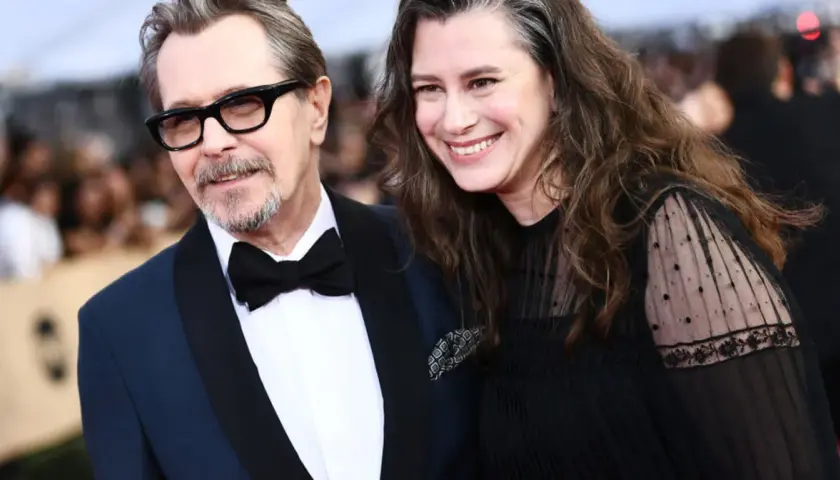 Gary Oldman z żoną, oboje uśmiechnięci, on w eleganckim garniturze, ona w czarnej sukni z koronkowymi rękawami.
