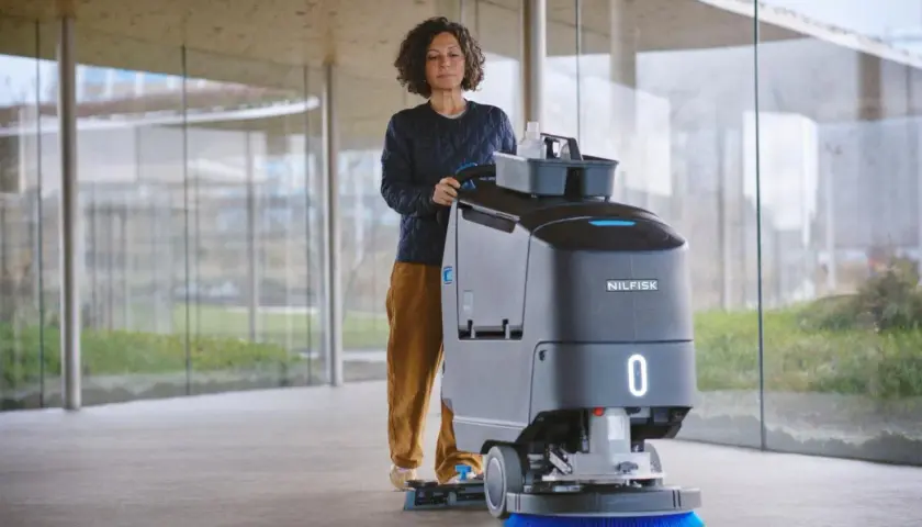 Mujer operando una máquina de Nilfisk, parte de sus avanzados sistemas de limpieza, en un pasillo moderno con grandes ventanales.
