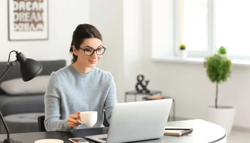 Kobieta w okularach pije kawę, pracując na laptopie przy biurku.
