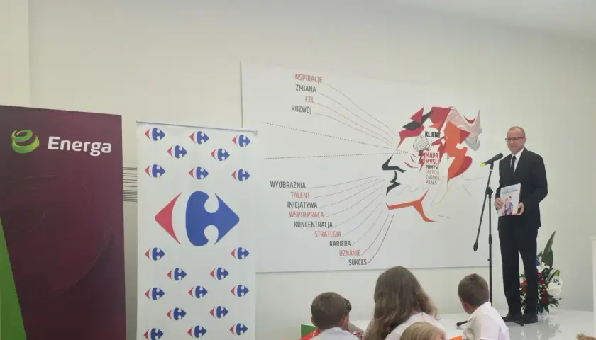 Mężczyzna w garniturze wręcza książkę "Karta Dużej Rodziny" na tle banerów Energa i Carrefour.