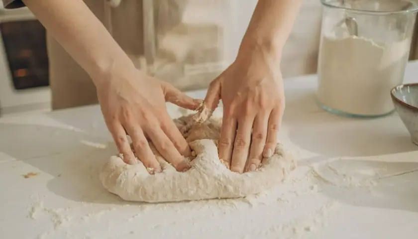 Manos amasando masa pan sobre una mesa con harina esparcida. Un frasco de vidrio con harina y un cuenco están al fondo.