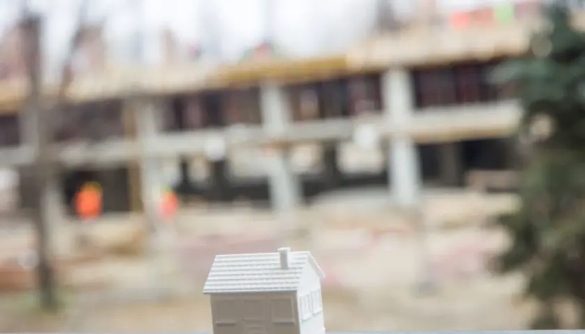 Biały model domu stoi na pierwszym planie, w tle widać budowę domu wielorodzinnego.