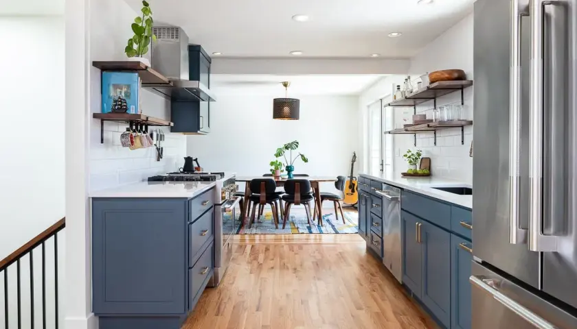 A modern kitchen with deep blue cabinets, white subway tile backsplash, and warm wood floors. The cabinet colors offer a stylish contrast to the stainless steel appliances.