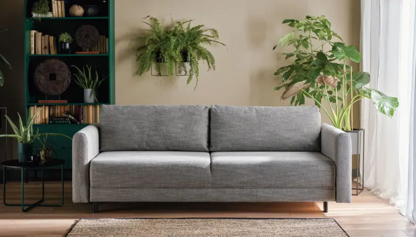 Sofa w kolorze popielaty jaki to kolor, otoczona zieloną biblioteczką i bujną roślinnością.