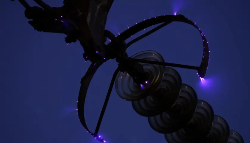 Des arcs électriques violets, les feux de Saint-Elme, crépitent sur une structure métallique et des isolateurs en verre contre un ciel nocturne.