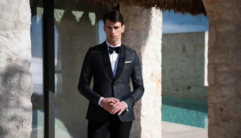 Elegancki mężczyzna w czarnym smokingu, gotowy na black tie event. Stoi przy basenie, otoczony kamiennymi murami.