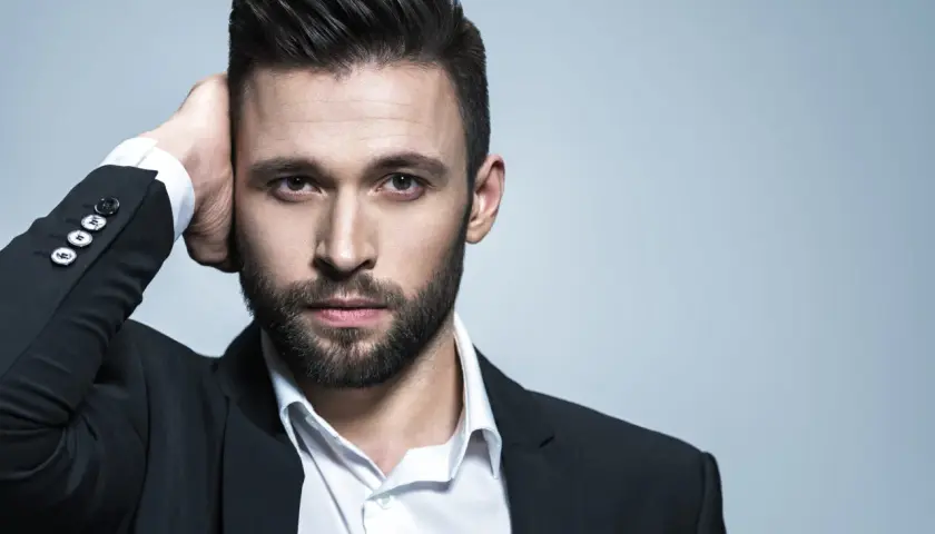 Hombre con un peinado con tupe, barba y traje negro, tocándose la cabeza.