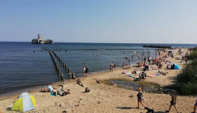 Słoneczna babie doły plaża tętni życiem. Ludzie opalają się, kąpią w morzu, a w oddali widać zabytkowy fort.