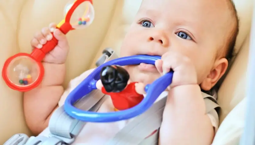Bambino con sonaglio rosso in mano e gioco blu in bocca, esplora il mondo con i sensi. Ottimo per stimolare neonato ad afferrare oggetti.