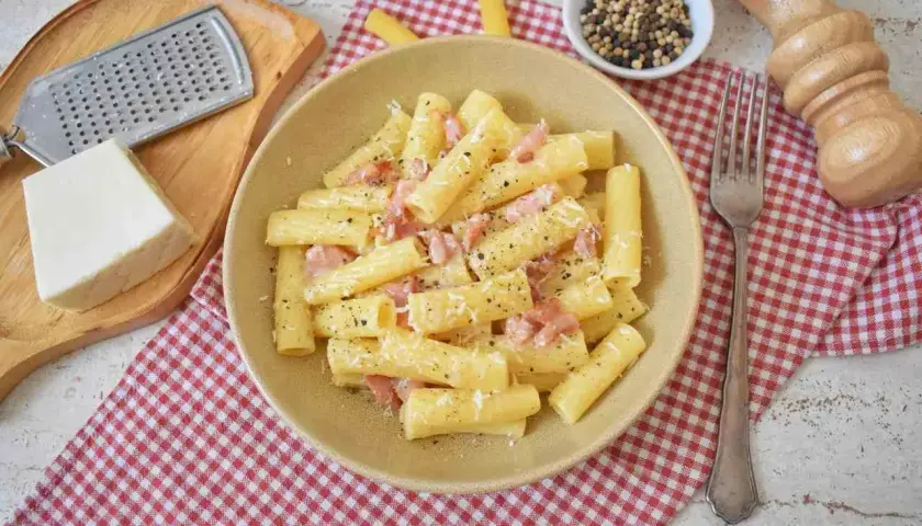 Pyszne rigatoni carbonara z boczkiem i parmezanem, posypane świeżo mielonym pieprzem. Obok tarka i kawałek sera.