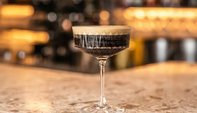 Un bicchiere di caffè martini con schiuma cremosa su un bancone di marmo, con luci sfocate sullo sfondo.
