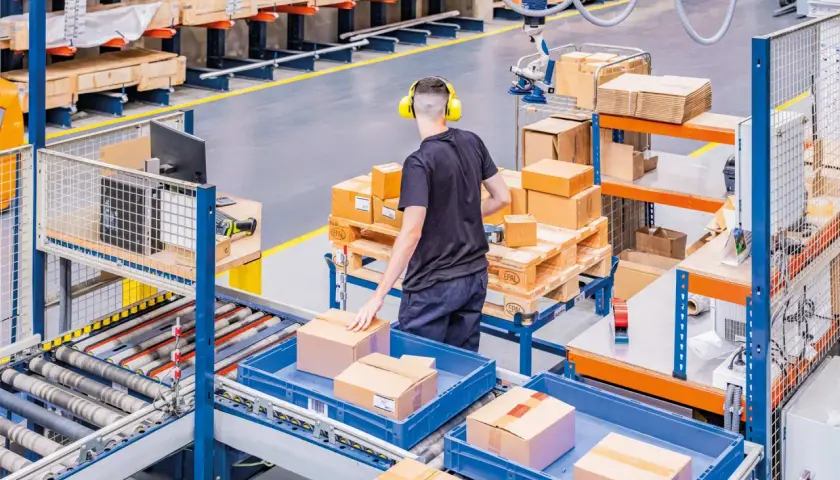 Ein Mitarbeiter mit Kopfhörern sortiert Pakete in einem Lager. Hier wird alles **assemble to order** für den Versand vorbereitet.