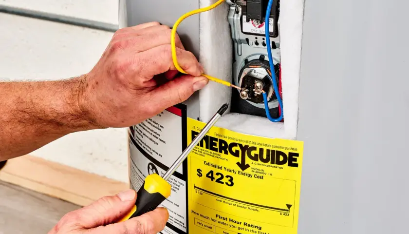 Ręce majsterkowicza podłączają przewody do bojlera, obok żółtej etykiety EnergyGuide, która informuje, ile prądu pobiera grzałka do bojlera.