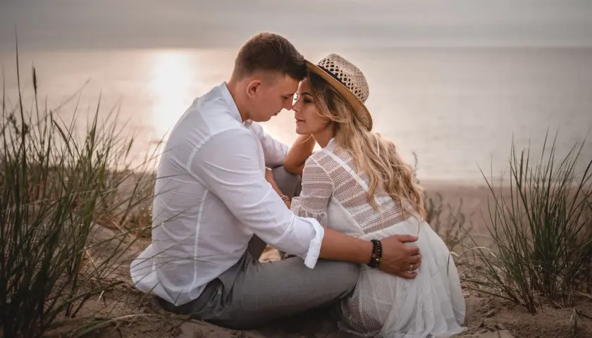 Romantyczna sesja we dwoje na plaży. Para siedzi blisko siebie, otoczona trawami, z widokiem na spokojne morze o zachodzie słońca.