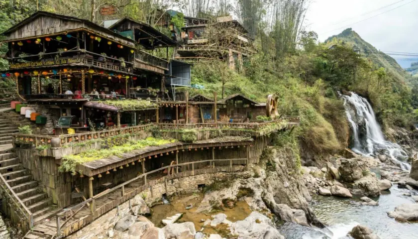 Holzgebäude schmiegen sich an einen Hang in Sapa, Vietnam, mit einem Wasserfall, der daneben herabstürzt.