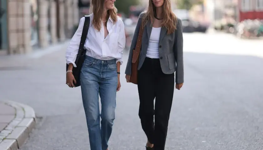 Dos mujeres caminan por la calle. Una, con un estilo demure, lleva vaqueros y camisa blanca. La otra, con chaqueta gris y pantalones negros, camina con elegancia.
