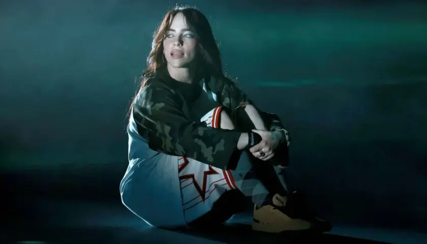 Billie Eilish siedzi na scenie, wpatrzona w dal. Jej stylizacja i mroczny klimat sugerują, że zaraz usłyszymy fajne piosenki.