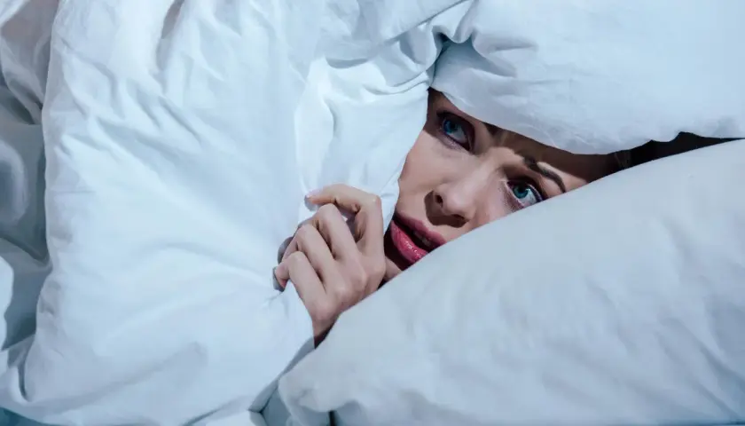 Verängstigte Frau versteckt sich unter der Decke, die Augen weit aufgerissen, als ob sie einen Albtraum erlebt.