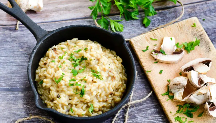 Risotto z grzybami i natką pietruszki w czarnej patelni, obok pokrojone pieczarki na desce.