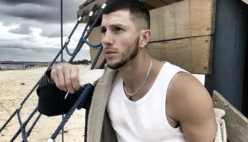 Sasha top model, z piórkiem w dłoni, na tle plaży i chmur.