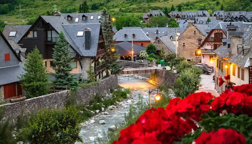 Pueblo pintoresco en el Pirineo Catalán, con casas de piedra y tejados grises junto a un río. Flores rojas en primer plano.
