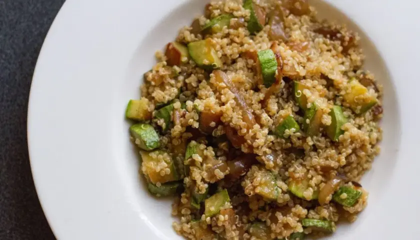 Un piatto di quinoa con verdure, tra cui zucchine a cubetti e cipolle caramellate, servito su un piatto bianco.