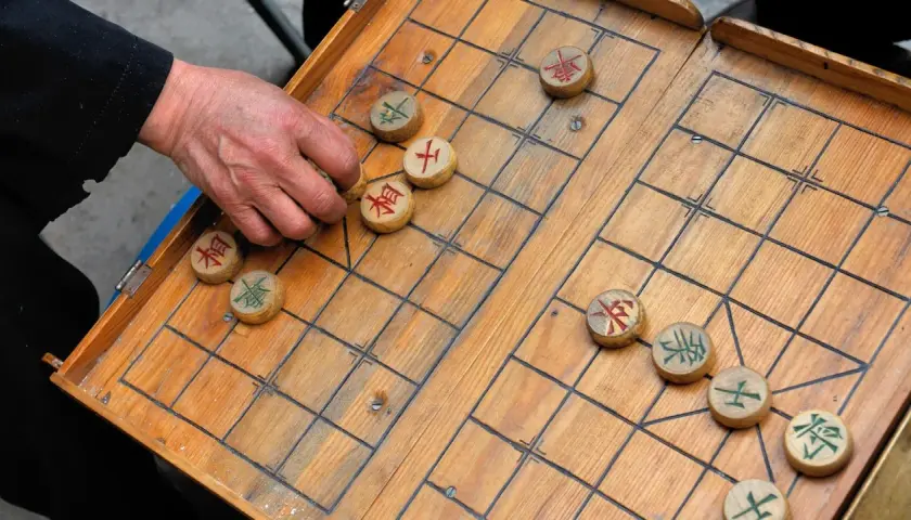 Eine Hand bewegt einen Stein beim China Schach. Das Holzbrett ist aufgeschlagen, die Figuren mit chinesischen Schriftzeichen sind verteilt.