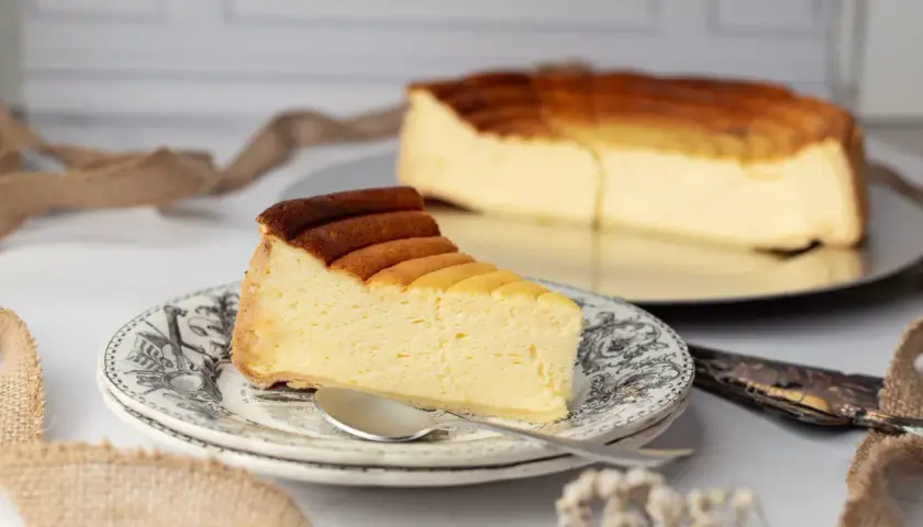 Une part de tarte au fromage, dorée et crémeuse, sur une assiette ornée. Le reste du gâteau est visible en arrière-plan.