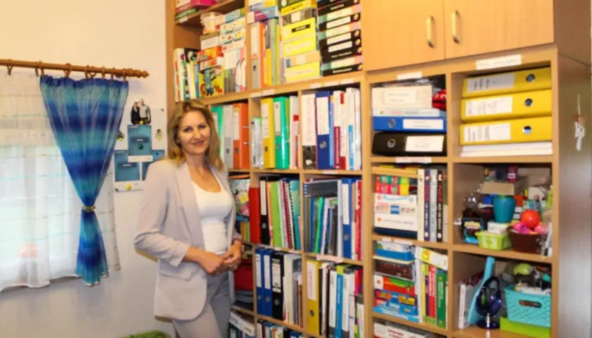 Kobieta w jasnej marynarce stoi w nowoczesnym gabinecie logopedycznym, otoczona regałami pełnymi materiałów terapeutycznych.