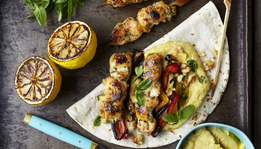 Grillad kyckling kebab med rostade grönsaker och hummus på ett tunnbröd, serverad med grillade citroner och mynta.