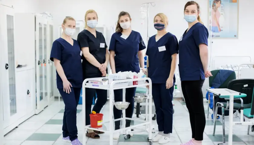 Grupa studentek pielęgniarstwa w maseczkach stoi przy wózku medycznym w sali szpitalnej.