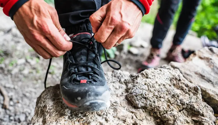 Dłonie zawiązują sznurówki czarnych butów podejściowych, gotowych na górską wyprawę. W tle widać inne buty trekkingowe.