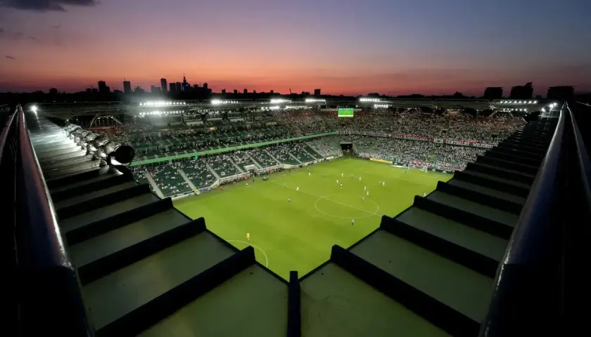 Zachód słońca nad stadionem Legii. Widok z góry na murawę, trybuny wypełnione kibicami i panoramę Warszawy. Pojemność stadionu Legii robi wrażenie.