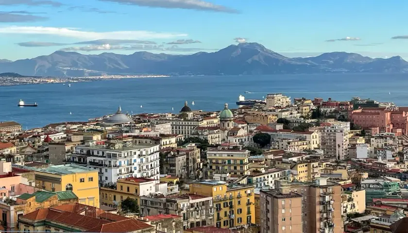 A panoramic view of Naples in December, with the blue sea and distant mountains under a cloudy sky.