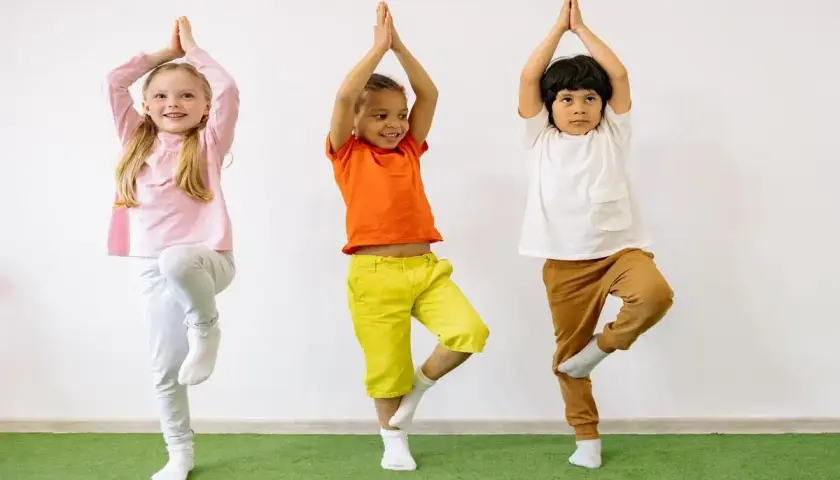 Tre bambini praticano yoga, in piedi su una gamba sola, con le mani giunte sopra la testa.
