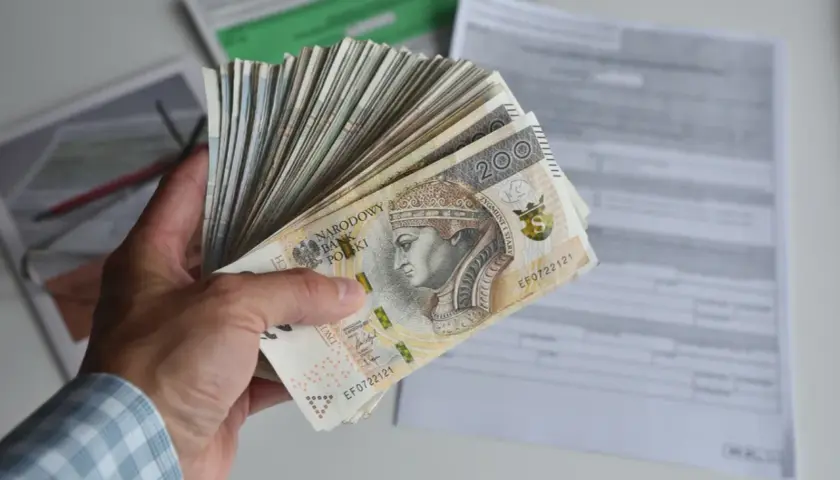 Dłoń trzyma plik banknotów 200 zł, symbolizujących dochód netto, na tle dokumentów podatkowych.