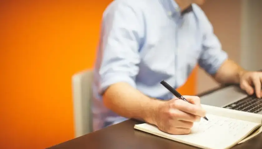 Mężczyzna w niebieskiej koszuli pisze notatki w zeszycie, pracując przy laptopie. Daje mu to **prokura** do działania.