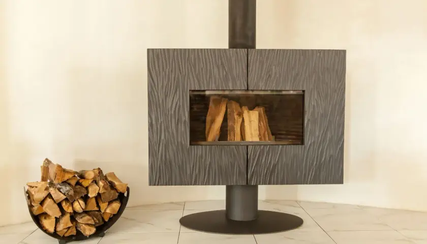 Un poêle moderne avec une cheminée vitrée et du bois de chauffage dans un panier. La **déco derrière un poêle** est sobre, mettant en valeur l'âtre.