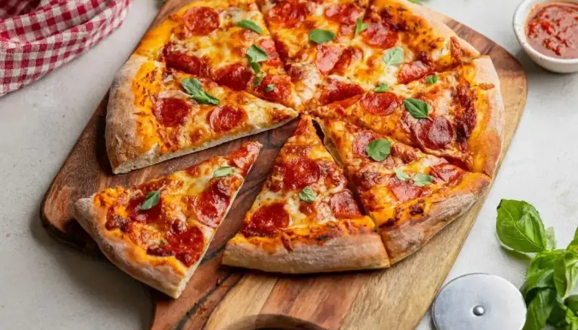 Pyszna pizza z pepperoni i świeżą bazylią, pokrojona na kawałki, leży na drewnianej desce. Obok sos pomidorowy i nóż do pizzy.