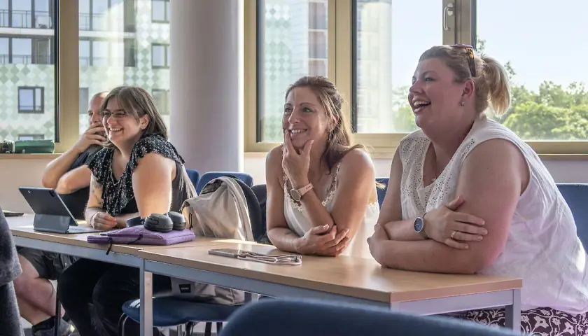 Lachende Teilnehmerinnen bei einem berufsbegleitenden Studium. Sie sitzen an Tischen mit Tablets und Notizbüchern.