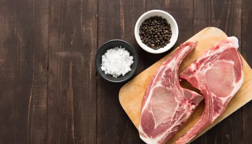 Deux belles côtes de porc crues sur une planche en bois, prêtes pour une cuisson au four, avec du sel et du poivre en grains à côté.