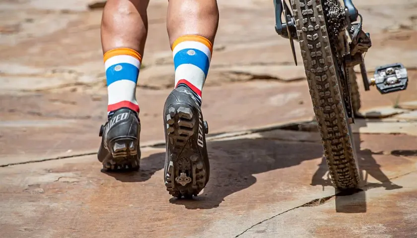 Mountain biking shoes with cleats are shown, not hiking boots. The rider's colorful socks and bike tire are also visible.