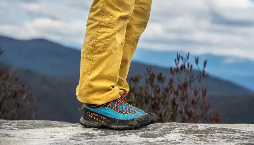 A person stands on a rock, wearing yellow pants and blue approach shoes with orange laces, ready for an adventure.