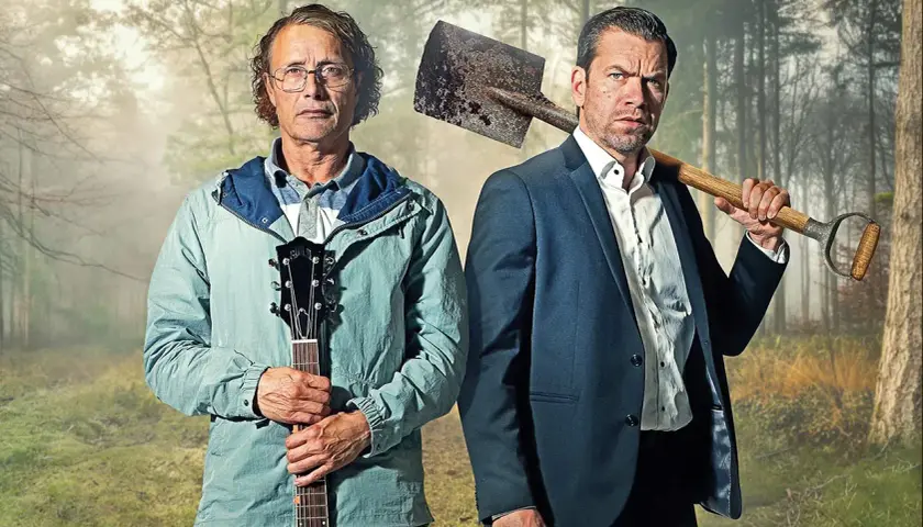 Två män i skogen, en med gitarr, en med spade. En scen från en mads mikkelsen serie.