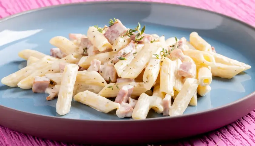 Penne pasta with creamy sauce and diced ham, garnished with thyme, on a blue plate.